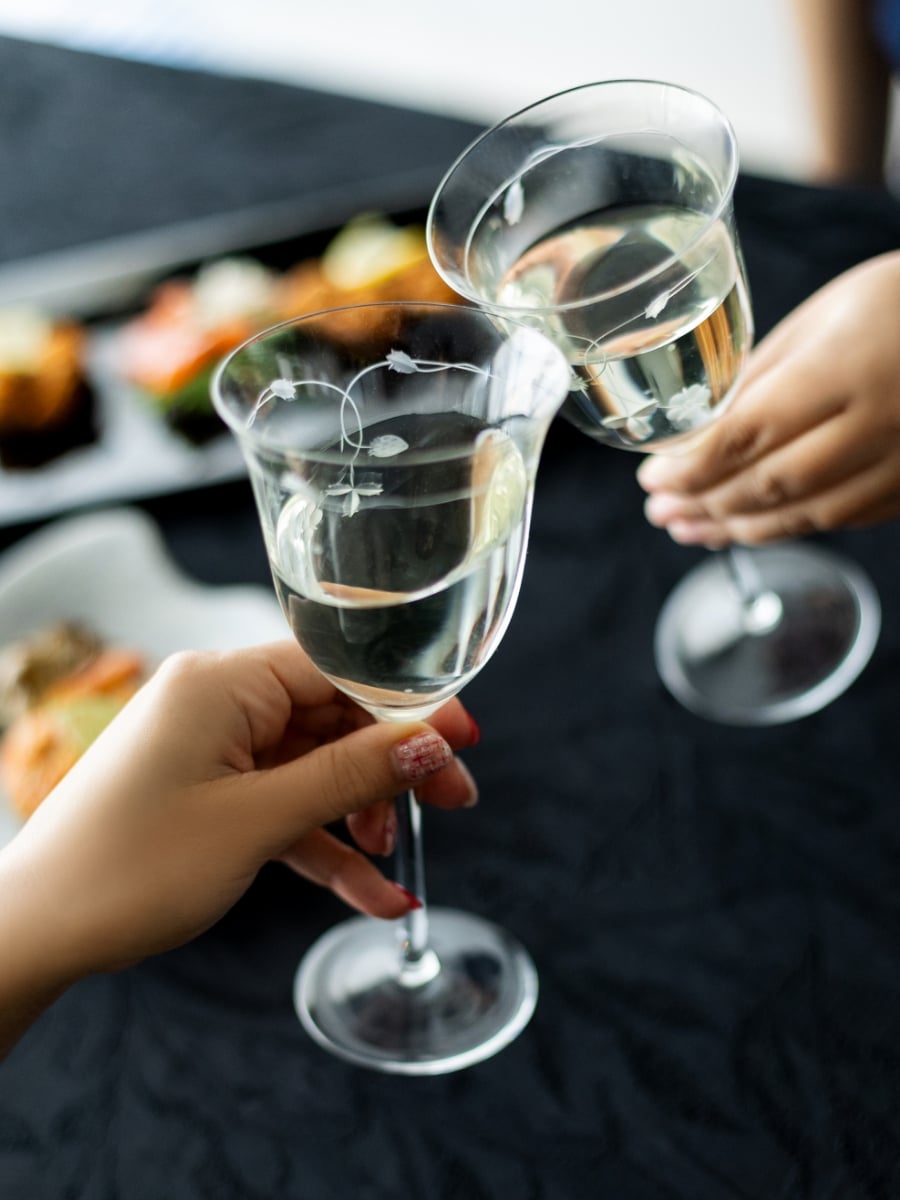 黒いテーブルの上で、二人が透明なグラスに入った白ワインを合わせて乾杯している場面。背景に和食の料理皿がぼんやりと見え、食事とともにワインを楽しむひとときを表している。