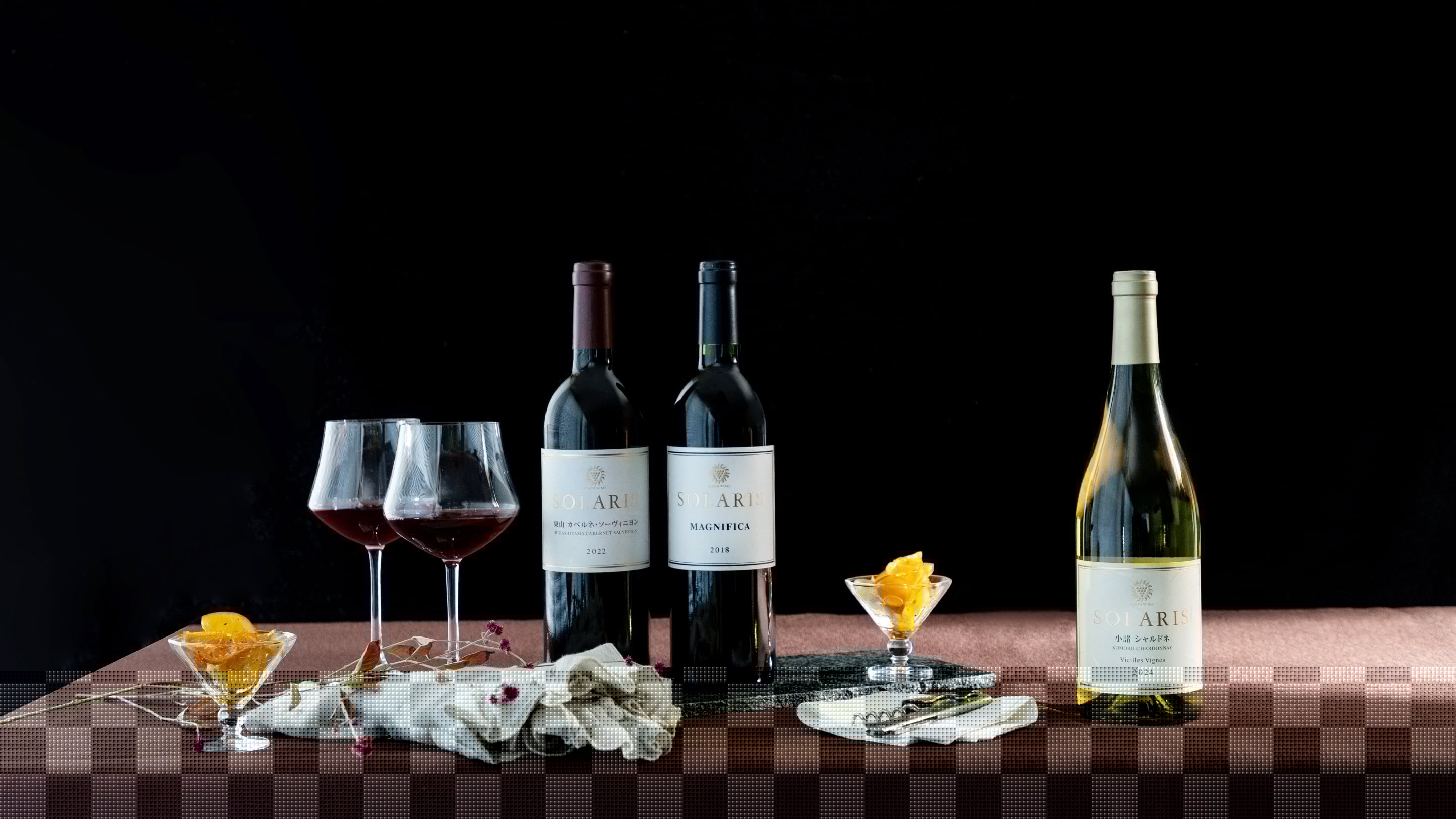 黒い背景の前に、SOLARISのワインを並べた静物写真。テーブルの上に赤ワイン2本と白ワイン1本、ワイングラス2脚、コルクスクリューとナプキン、小皿に盛られたドライフルーツが配置され、落ち着いた高級感のある雰囲気
