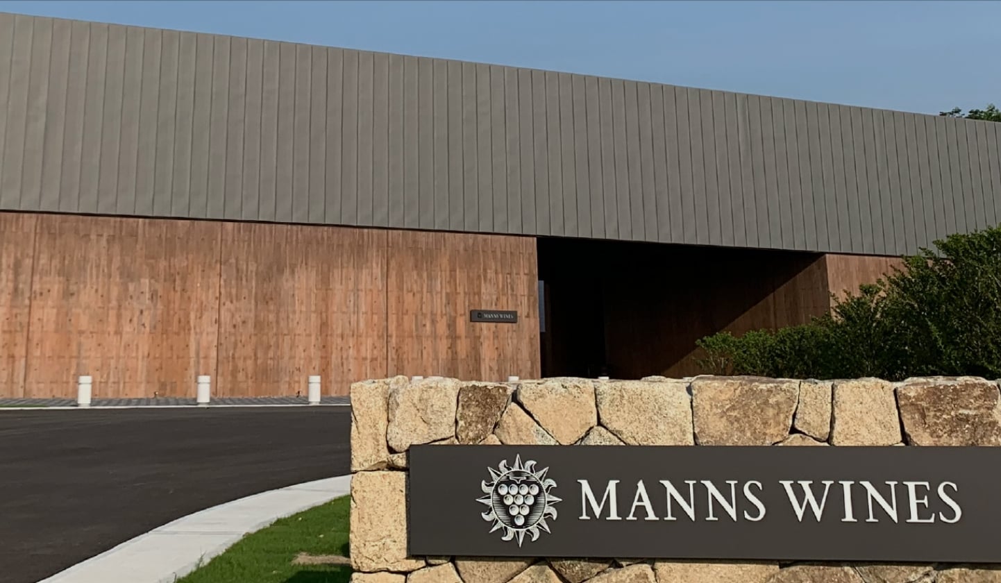 石積みのサインに『MANNS WINES』と表示された、マンズワインの勝沼ワイナリー入口。奥に木材を使った建物があり、敷地内へ続くアプローチと建物外観が写っている。
